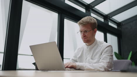 handsome-man-is-working-with-laptop-in-modern-office-in-daytime-person-in-white-shirt-is-surfing-internet